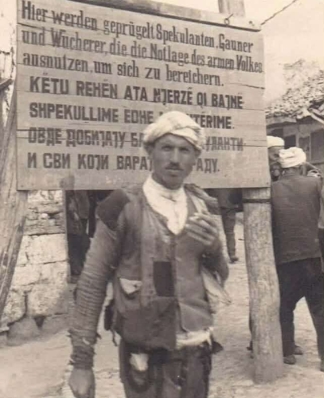 <br />
Në ballë të kësaj fotografie rri një shqiptar i moshës së mesme, i veshur me rroba të arrnuara me lecka e me tjerq galana, pantollona tradicional leshi për burra me ngjyrë të kafenjtë dhe shitira gajtani të zi, teksa me një cigare duhani në dorë, pozon në drejtim të objektivit fotografik. Më shumë sesa te vetë ai, vëmendja shkon mbrapa tij te një tabelë druri në sfond, në të cilën afërsisht i njëjti njoftim paralajmërues është shkruar në gjermanisht, shqip dhe serbisht. Teksti në gjermanisht ka këtë përmbajtje: “Këtu rrihen spekulantët dhe mashtruesit, si dhe fajdexhinjtë që shfrytëzojnë gjendjen e vështirë të njerëzve të varfër për t’u pasuruar”. Poshtë këtij teksti, në dy rreshta me shkronja të mëdha në shqip është shkruar: “Këtu rehën ata njerëz qi bajnë shpekullime dhe mashtërime”. 