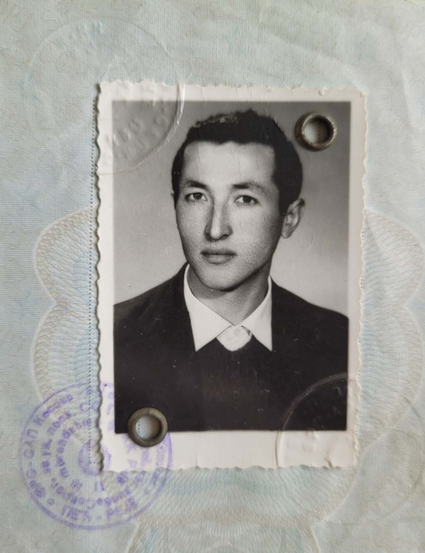 Fotografi bardh e zi, e ngjitur në pasaportë. Portret i një burri të ri, me flokë të shkurtëra, veshur me kostume të zeza dhe këmishë të bardhë.