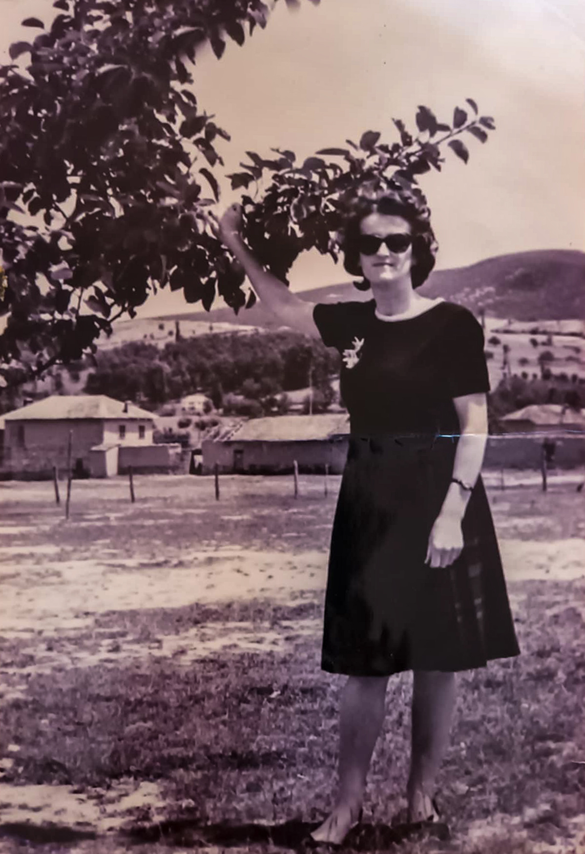 The image is a black and white photograph of a woman standing outdoors. She is wearing a dark dress and sunglasses, and her hair is styled in a short, wavy fashion. The woman is smiling and has one arm raised, touching the branches of a tree beside her. In the background, there are hills and some buildings, suggesting a rural or suburban setting.