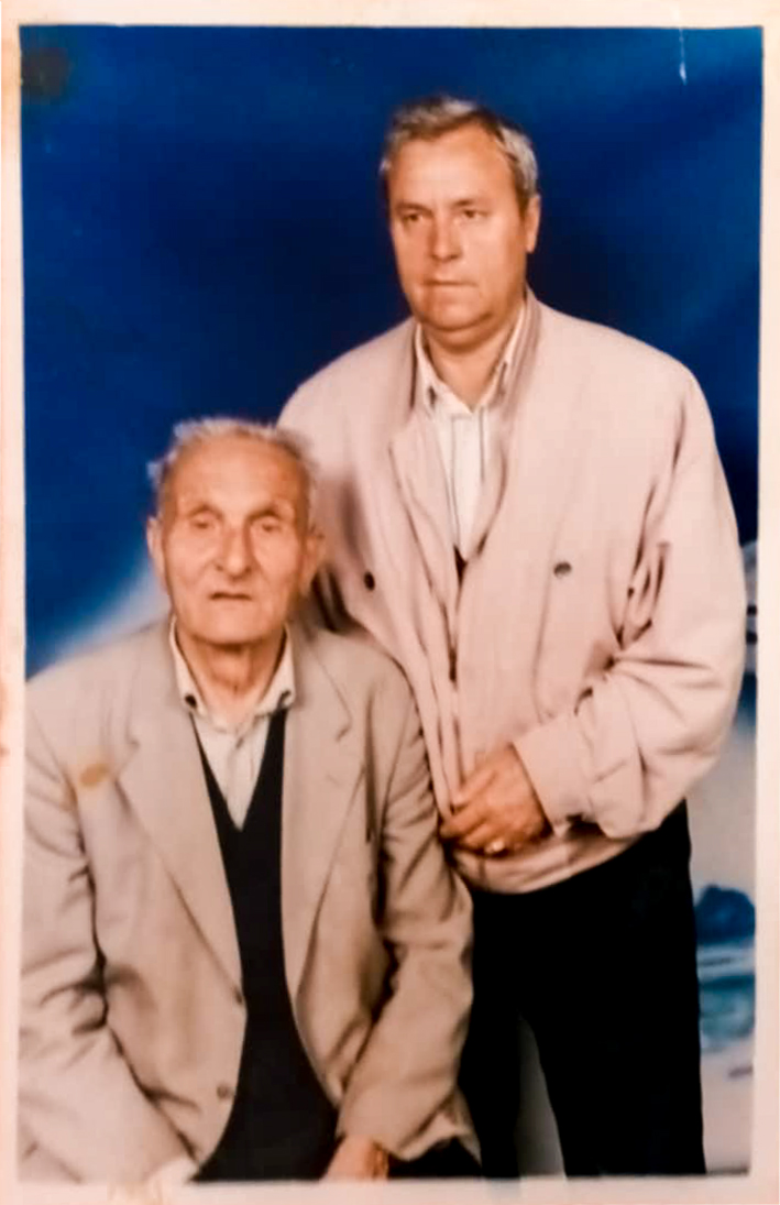 The image shows a photograph of two men. The man on the left is seated and appears to be older, with gray hair, wearing a light-colored suit jacket over a dark sweater and a light shirt. The man on the right is standing, with his hand resting on the seated man's shoulder. He has short gray hair and is wearing a light-colored jacket over a light shirt. The background of the photograph is a solid blue color.