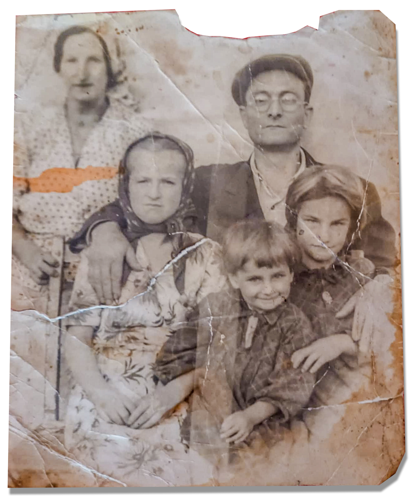 The image shows an old, sepia-toned photograph of a family. The photo is worn and has visible creases and a torn section at the top. It features five people: a man, a woman, and three children. The man is wearing a cap and glasses, and the woman is standing behind him. The children are positioned in front of the adults, with one child sitting on the lap of the woman. The photograph is placed on a pink textured surface, possibly a cloth or paper.