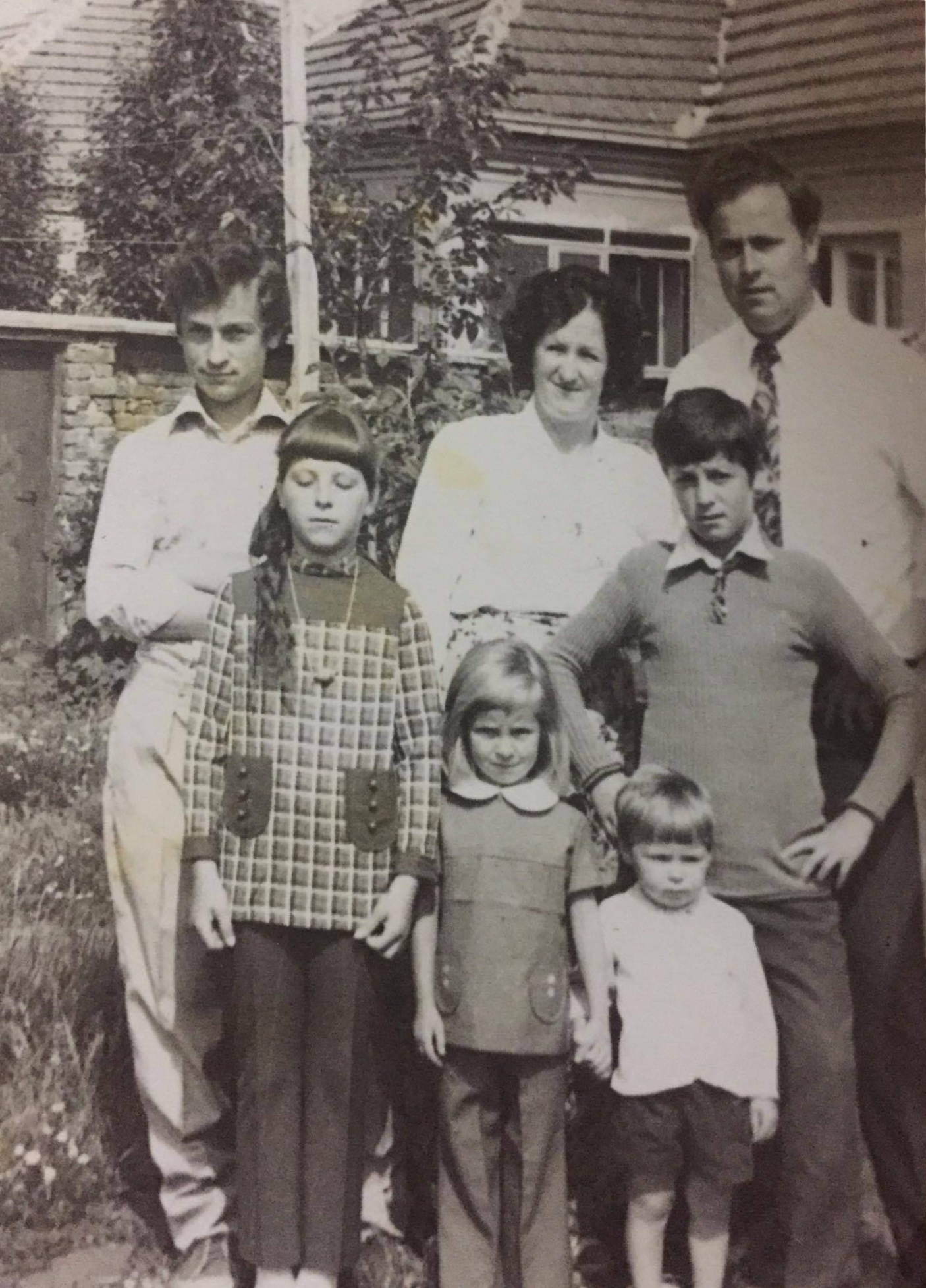 This is a black and white photograph of a family group. There are seven people in the picture, standing in front of a house with a tiled roof and some foliage in the background. In the back row, there are three adults. On the left is a man with short hair, wearing a light-colored shirt and trousers. In the middle is a woman with dark hair, wearing a light-colored blouse. On the right is another man with short hair, wearing a light-colored shirt and a tie. In the front row, there are four children. On the left is a girl with short hair, wearing a patterned dress with a collar and buttons. Next to her is another girl with glasses and a similar dress. To the right of her is a boy with short hair, wearing a sweater and trousers. On the far right is a young child, possibly a toddler, wearing a light-colored top and dark shorts.