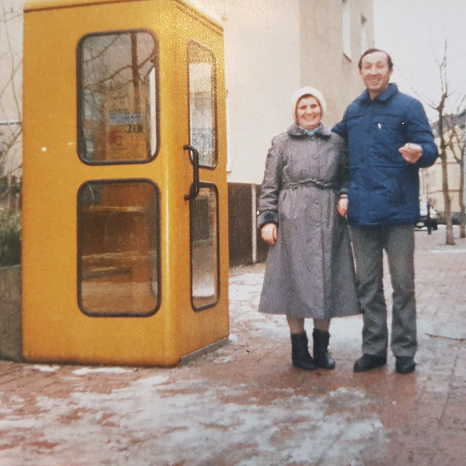 A man and a woman stand next to each other. The woman wears a gray coat and a headscarf, while the man wears a blue jacket. A yellow phone booth stands near them.