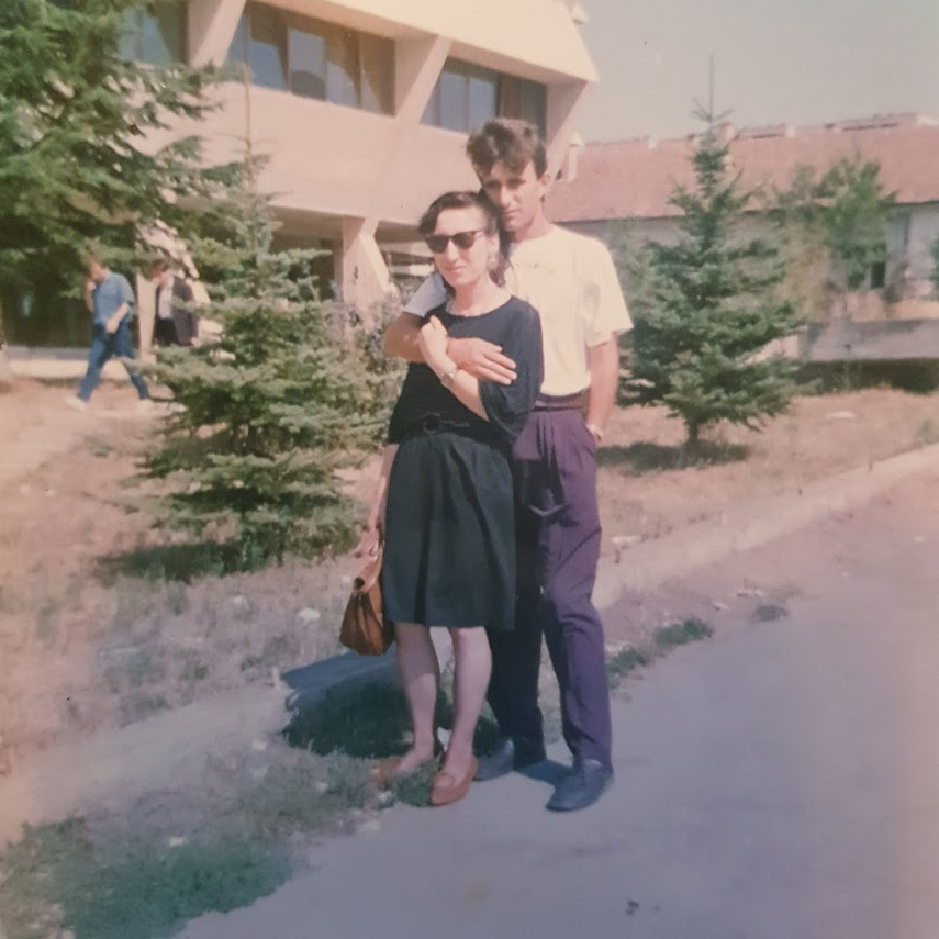 A man and a woman stand together. The woman, in front, wears a dress and sunglasses, while the man stands behind her, hugging her with one arm. He wears a white T-shirt and dark-colored pants.