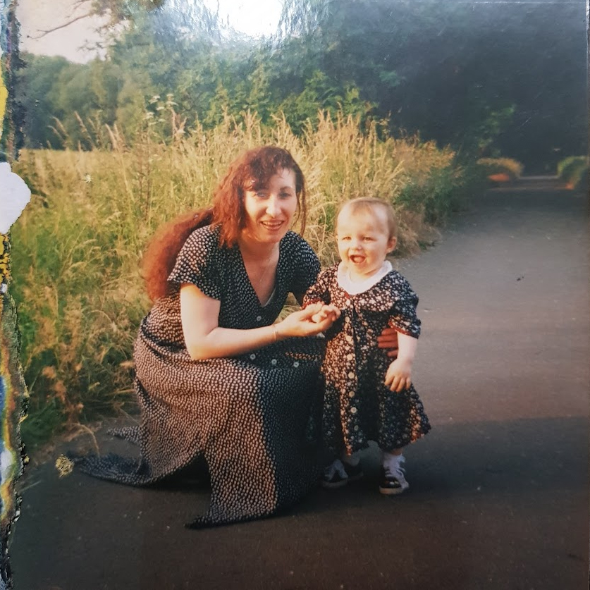 A woman and a child sit on a road surrounded by grass and trees. The woman, on the left, smiles while wearing a black dress with white dots. The child, on the right, also smiles, wearing a similar dress.